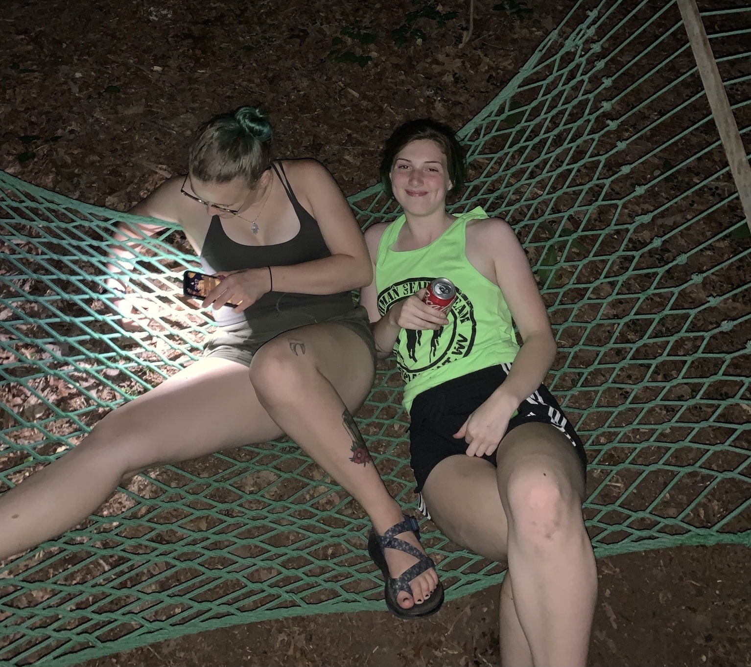 JQ and Ashley sitting in a green hammock. JQ has her phone flashlight on shinning through the hammock holes.